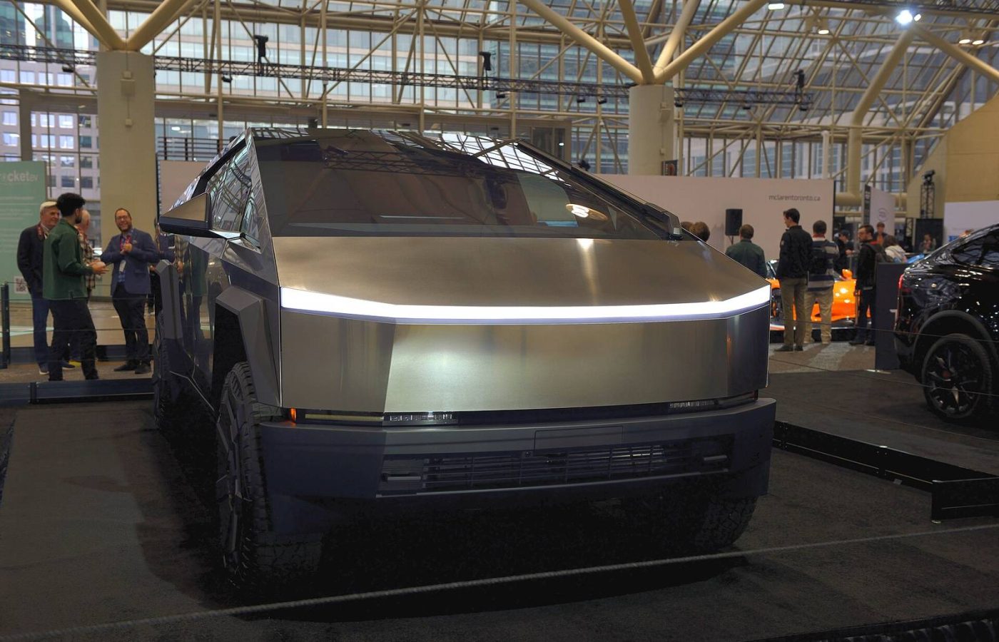 Front view of a stainless steel Tesla Cybertruck with its light bar on, displayed indoors