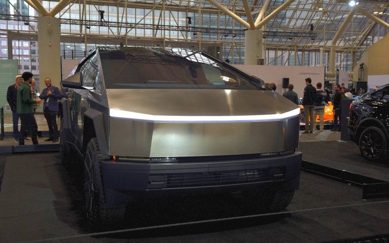Front view of a stainless steel Tesla Cybertruck with its light bar on, displayed indoors