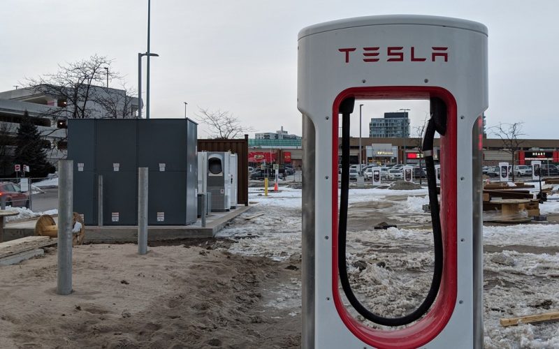 White and red Tesla Supercharger post in a parking lot with patches of snow and a shopping mall behind