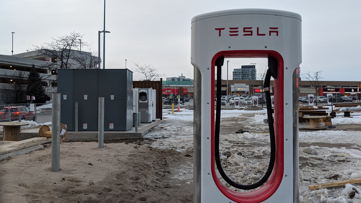 White and red Tesla Supercharger post in a parking lot with patches of snow and a shopping mall behind