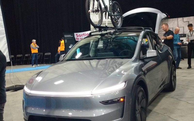 Silver 2025 Tesla Model Y with its rear liftgate open and a bike on the roof at an indoor auto show
