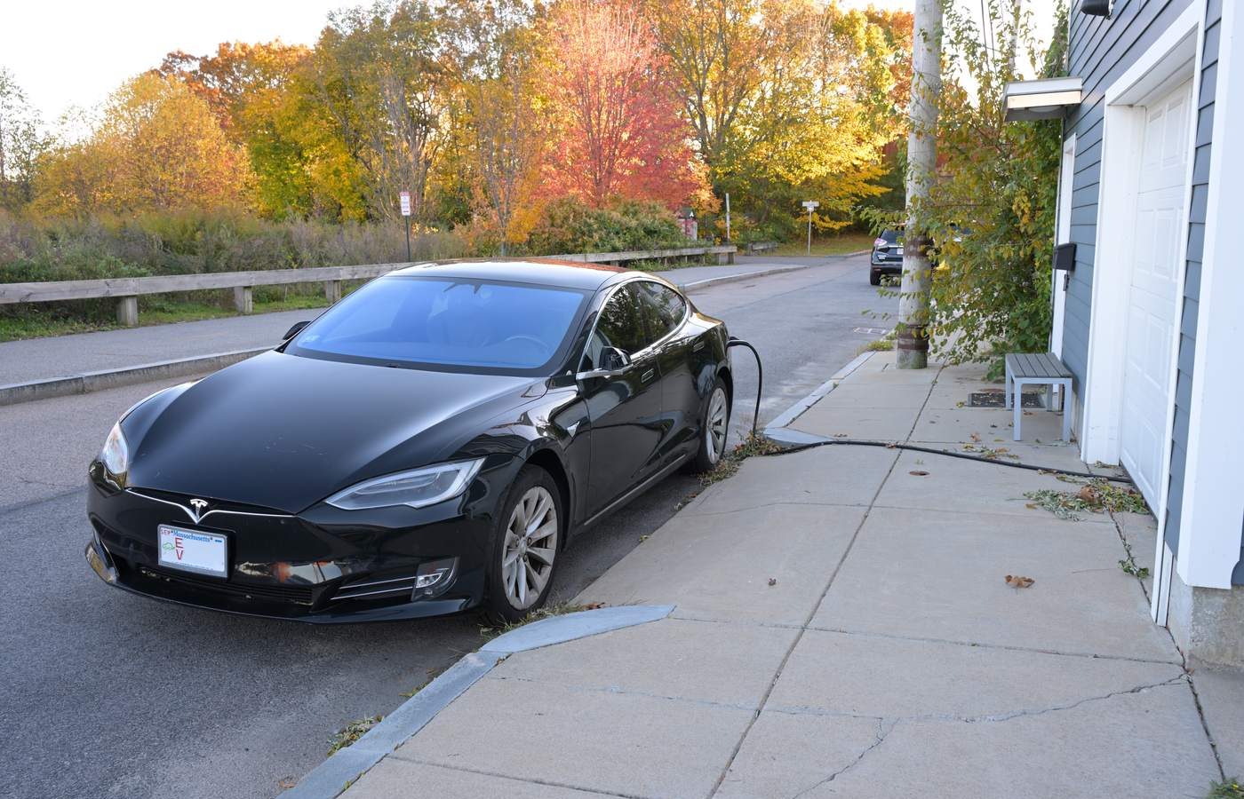 Black Tesla Model S plugged into a home charging cable running from a garage, with autumn trees behind it.