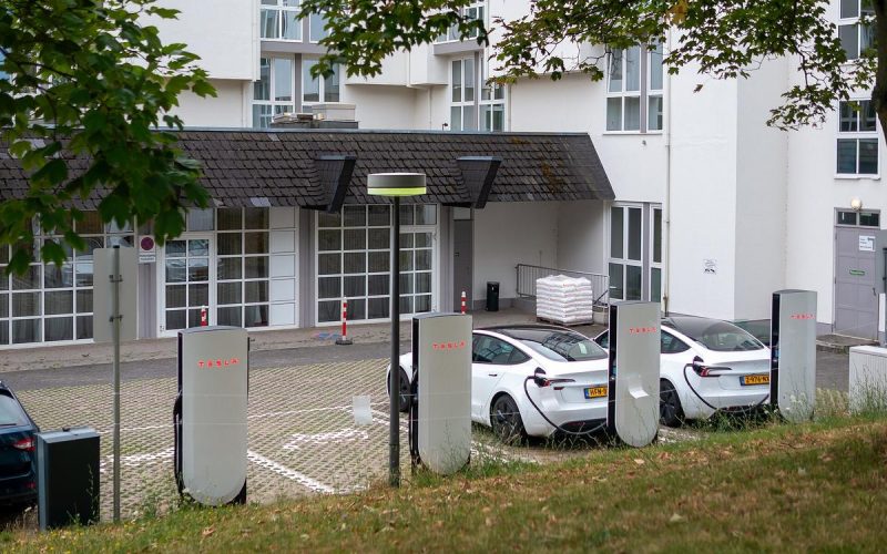 Row of white Tesla V4 Supercharger posts with two white Tesla cars plugged in beside a building