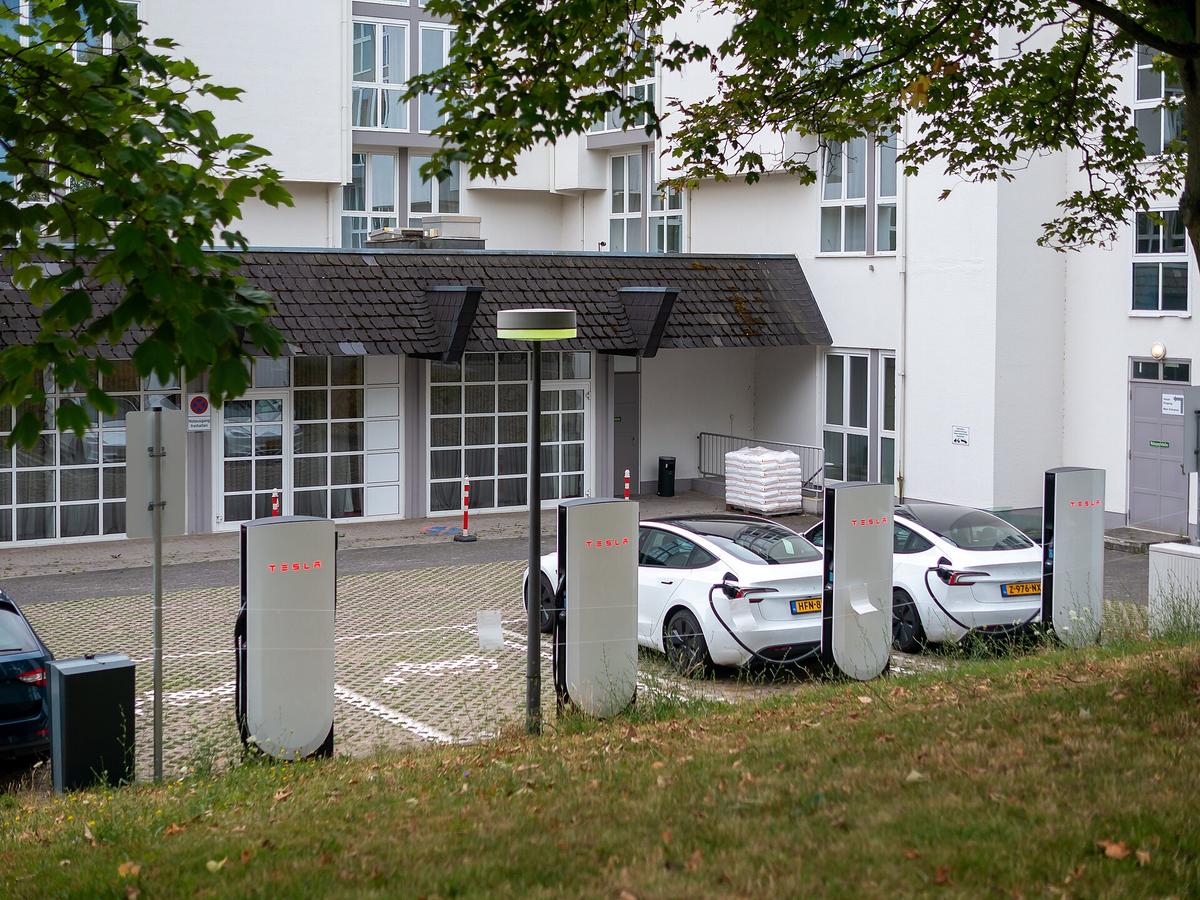 Row of white Tesla V4 Supercharger posts with two white Tesla cars plugged in beside a building