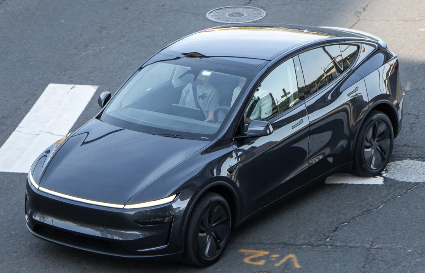 Dark grey new-generation Tesla Model Y with a full-width front light bar driving through a city intersection.