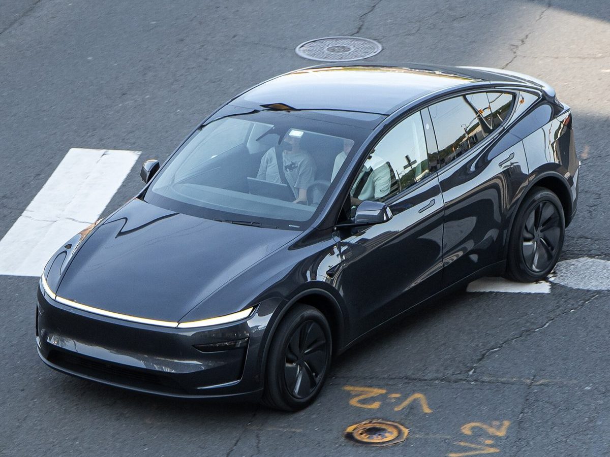 Overhead view of a dark grey refreshed Tesla Model Y with a full-width front light bar crossing a street