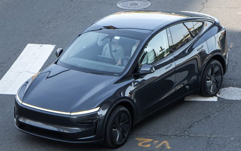 Overhead view of a dark grey refreshed Tesla Model Y with a full-width front light bar crossing a street