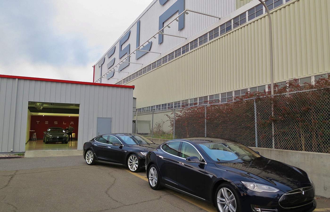 Two dark blue Tesla Model S sedans parked outside Tesla's Fremont factory under a large TESLA sign