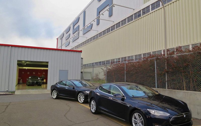 Two dark blue Tesla Model S sedans parked outside Tesla's Fremont factory under a large TESLA sign