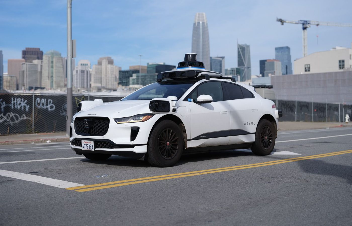 White Waymo Jaguar I-Pace robotaxi with roof-mounted sensors on a city street, downtown skyline behind it