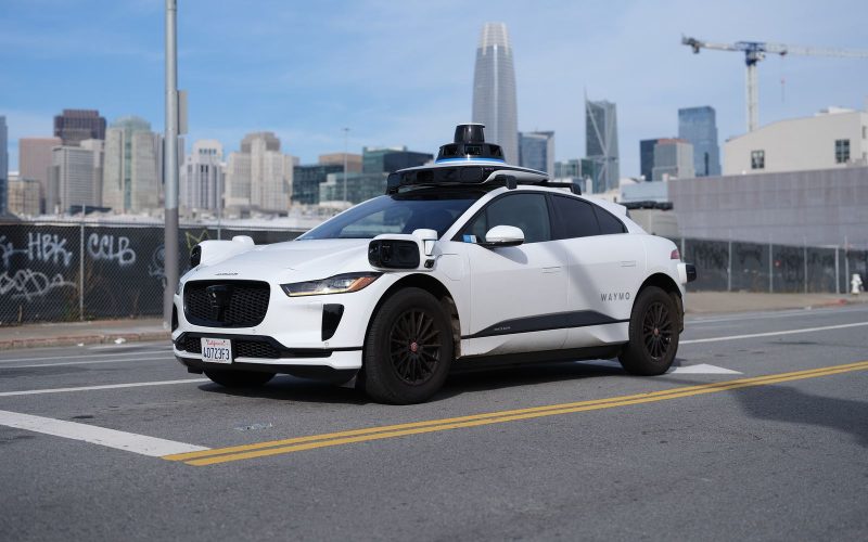 White Waymo Jaguar I-Pace robotaxi with roof-mounted sensors on a city street, downtown skyline behind it